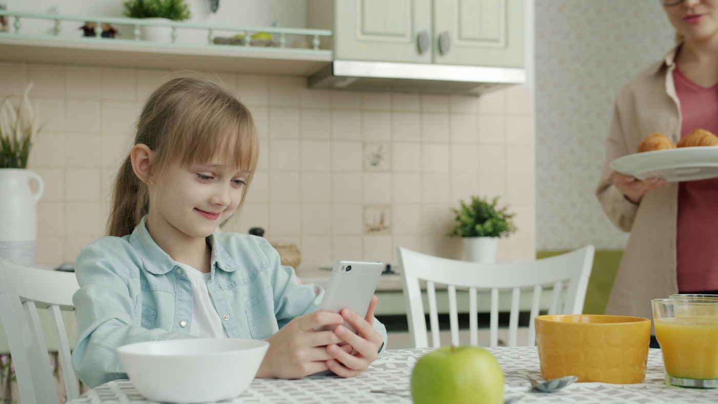 Young girl using a smartphone at the kitchen table while her mother brings breakfast — the family's safety scavenger hunt in action